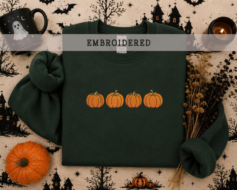 May include: A dark forest green crewneck sweatshirt featuring an embroidered design of four orange pumpkins in a horizontal row across the center. The garment has a ribbed collar, cuffs, and hem, with the word EMBROIDERED printed in a gray bar.