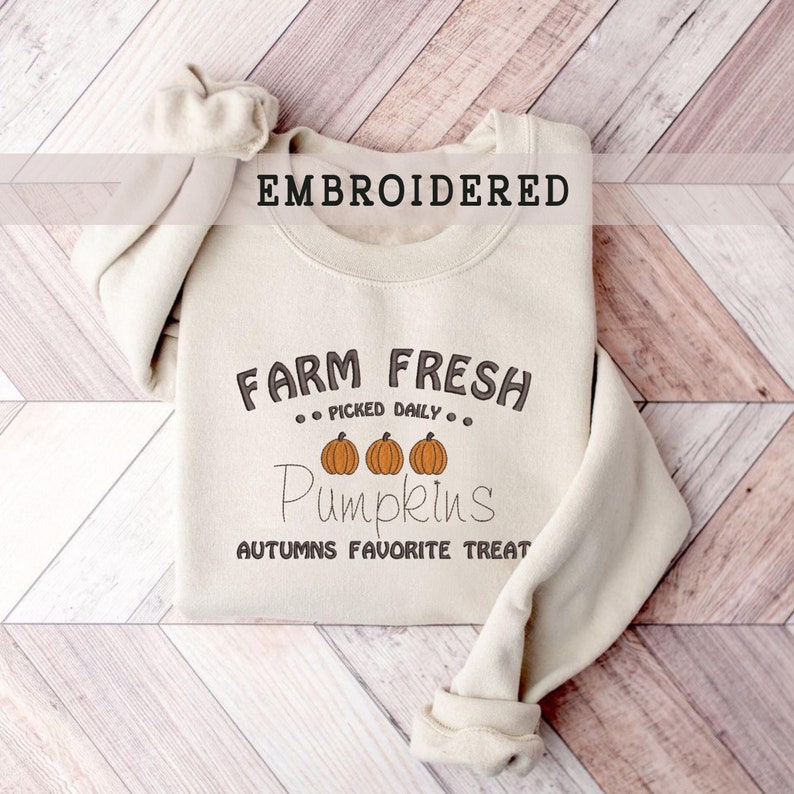 Op de afbeelding: Een cr&egrave;mekleurige sweatshirt met het woord "EMBROIDERED" bovenaan. De sweatshirt heeft de tekst "FARM FRESH PICKED DAILY" boven drie oranje pompoenillustraties en het woord "Pumpkins" in een scriptlettertype. De zin "AUTUMNS FAVORITE TREAT" staat eronder.