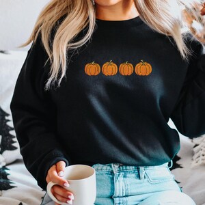 May include: A black long-sleeve crewneck sweatshirt featuring an embroidered design of four orange pumpkins with green stems arranged in a horizontal row across the center of the chest. The garment has ribbed cuffs and a ribbed neckline.