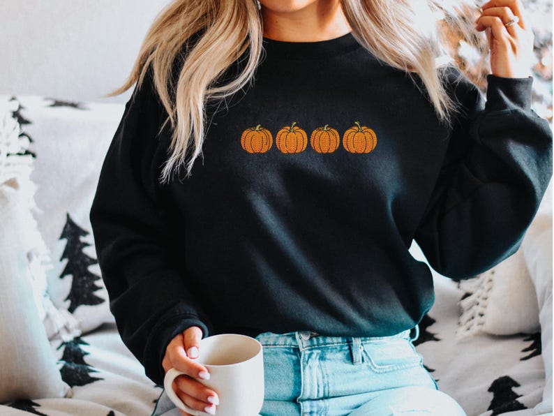 May include: A black long-sleeve crewneck sweatshirt featuring an embroidered design of four orange pumpkins with green stems arranged in a horizontal row across the center of the chest. The garment has ribbed cuffs and a ribbed neckline.