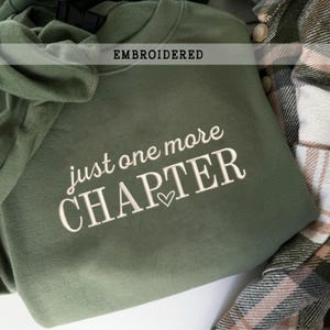 Pode incluir: Camisola verde-oliva com a frase bordada "just one more CHAPTER" em escrita branca, com um pequeno s&iacute;mbolo de cora&ccedil;&atilde;o. A palavra "EMBROIDERED" est&aacute; impressa num ret&acirc;ngulo branco acima do texto. Uma camisa xadrez &eacute; vis&iacute;vel.