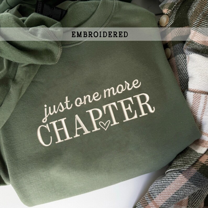 Pode incluir: Camisola verde-oliva com a frase bordada "just one more CHAPTER" em escrita branca, com um pequeno s&iacute;mbolo de cora&ccedil;&atilde;o. A palavra "EMBROIDERED" est&aacute; impressa num ret&acirc;ngulo branco acima do texto. Uma camisa xadrez &eacute; vis&iacute;vel.