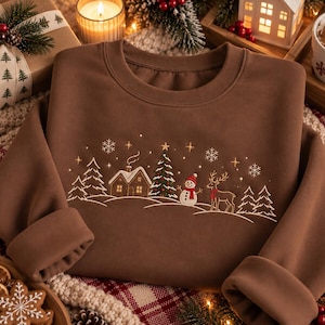 Könnte beinhalten: Ein braunes Sweatshirt mit Rundhalsausschnitt und einer gestickten Winterszene. Das Design zeigt eine kleine Hütte, einen geschmückten Weihnachtsbaum, einen Schneemann mit rotem Schal und ein Rentier auf einem verschneiten Hügel.