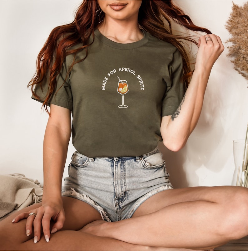 Puede incluir: Una mujer con una camiseta verde con el texto "Made for Aperol Spritz" y un vaso de Aperol Spritz. Est&aacute; sentada con las piernas cruzadas.