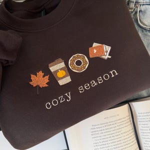 Peut inclure: Un sweat-shirt marron foncé avec la phrase brodée "cozy season" en blanc. Le motif comprend une feuille d'automne, une tasse de café avec une citrouille, un beignet et une petite pile de livres.