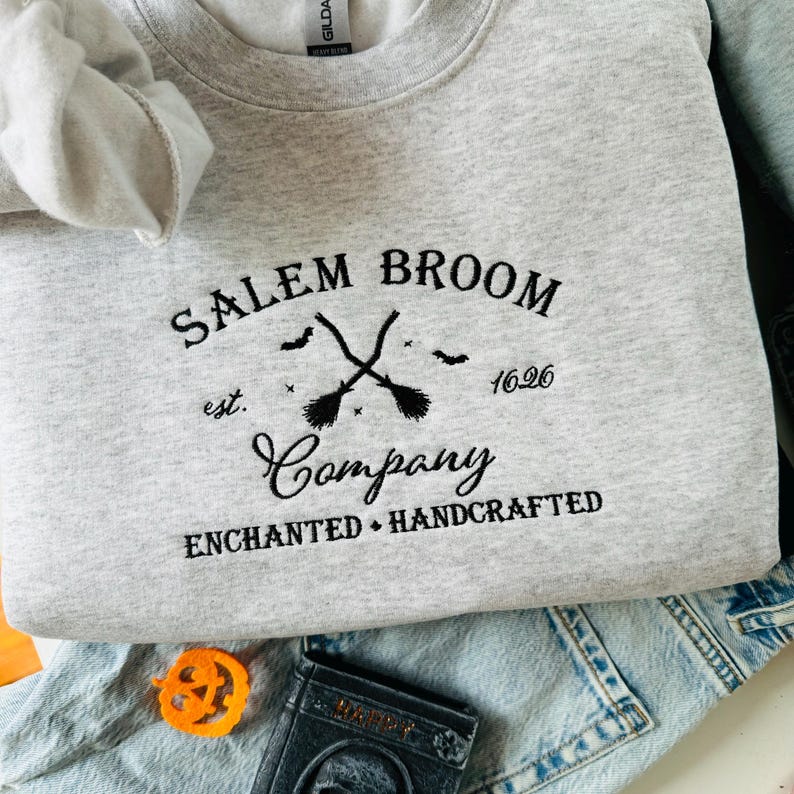 K&ouml;nnte beinhalten: Ein graues Sweatshirt mit einem schwarzen gestickten Design, das "Salem Broom Company est. 1626 Enchanted Handcrafted" lautet. Das Design zeigt zwei gekreuzte Besen mit Sternen und Flederm&auml;usen.