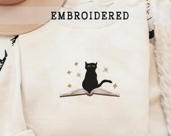 以下が含まれることがあります： 前面に「EMBROIDERED」という文字がプリントされた白いスウェットシャツ。スウェットシャツには、開いた本の上に座っている黒い猫と、その周りを囲む金色の星が刺繍されています。