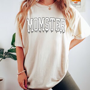 May include: A white t-shirt with the text "MOMSTER" in black dripping letters. The shirt is shown on a model and is labeled "IVORY".