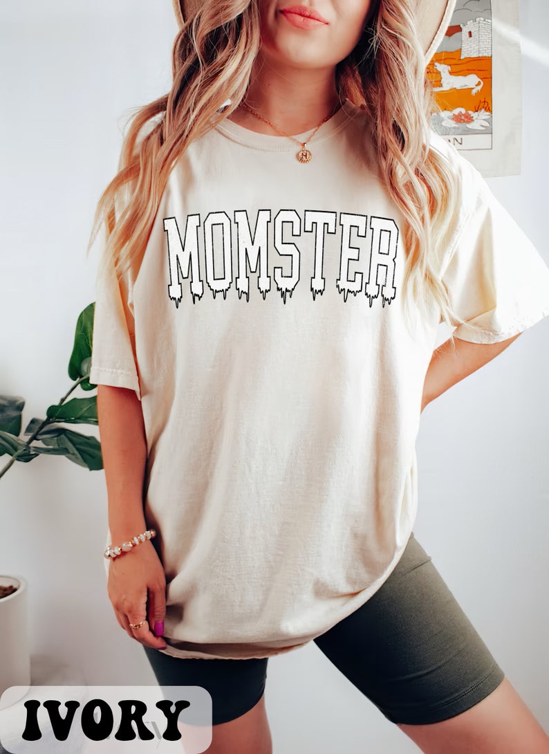 May include: A white t-shirt with the text "MOMSTER" in black dripping letters. The shirt is shown on a model and is labeled "IVORY".