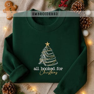 Könnte beinhalten: Dunkelgrünes Sweatshirt mit gesticktem Weihnachtsbaum-Design. Der Baum besteht aus Büchern und Lichterketten, gekrönt von einem goldenen Stern. Der Text "all booked for Christmas" steht darunter. Das Wort "EMBROIDERED" steht oben.