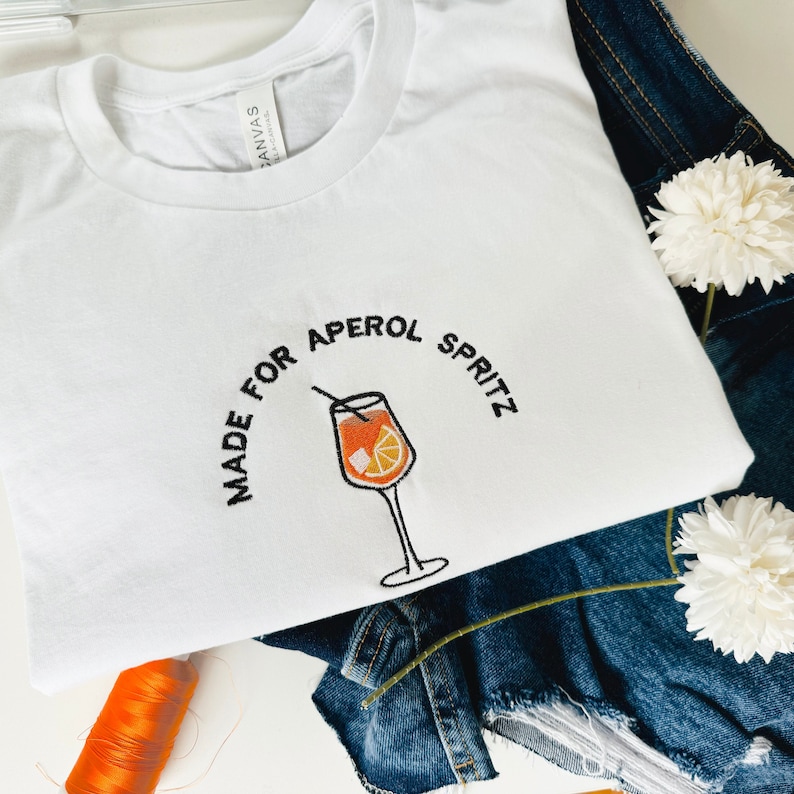 Puede incluir: Camiseta blanca con bordado negro que dice "Made for Aperol Spritz" con una ilustraci&oacute;n de un c&oacute;ctel Aperol Spritz.