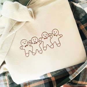 Może przedstawiać: Kremowa bluza z haftowanym wzorem czterech piernikowych ludzików trzymających się za ręce. Piernikowe ludziki są brązowe i mają brązowe oczy i uśmiechy.