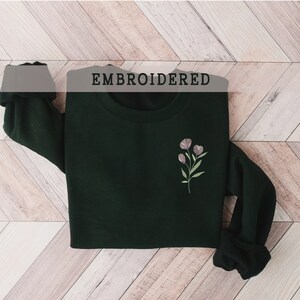 May include: A dark green sweatshirt with a small embroidered floral design on the left chest. The text "EMBROIDERED" is printed above the sweatshirt.