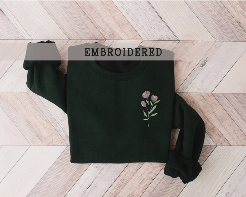 May include: A dark green sweatshirt with a small embroidered floral design on the left chest. The text "EMBROIDERED" is printed above the sweatshirt.