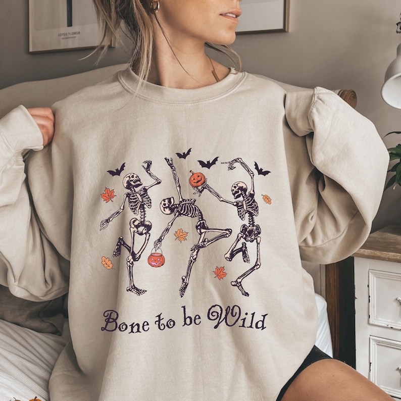 K&ouml;nnte beinhalten: Ein beiger Pullover mit einem Aufdruck von drei tanzenden Skeletten, die K&uuml;rbisse halten. Der Text "Bone to be Wild" ist unter den Skeletten aufgedruckt.