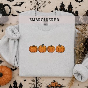 May include: A light gray crewneck sweatshirt featuring an embroidered design of four orange pumpkins in a horizontal row. The word EMBROIDERED is printed in black capital letters at the top center of the garment, positioned above the neckline.