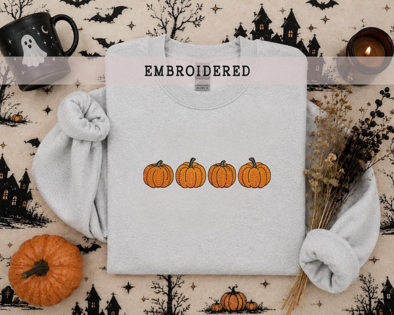 May include: A light gray crewneck sweatshirt featuring an embroidered design of four orange pumpkins in a horizontal row. The word EMBROIDERED is printed in black capital letters at the top center of the garment, positioned above the neckline.