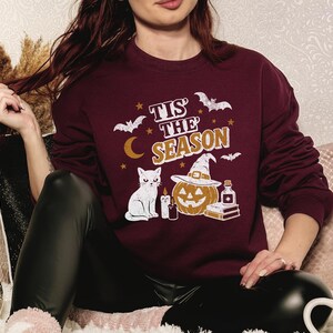 May include: A maroon sweatshirt with a Halloween-themed graphic. The design includes the text "Tis the Season", bats, a crescent moon, stars, a white cat, a jack-o'-lantern wearing a witch hat, candles, and a potion bottle. The sweatshirt is long-sleeved.