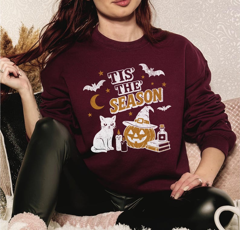 May include: A maroon sweatshirt with a Halloween-themed graphic. The design includes the text "Tis the Season", bats, a crescent moon, stars, a white cat, a jack-o'-lantern wearing a witch hat, candles, and a potion bottle. The sweatshirt is long-sleeved.