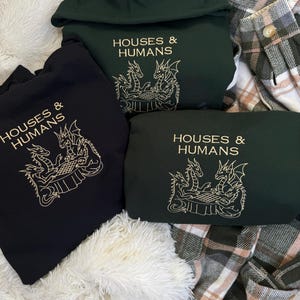 May include: Three hooded sweatshirts in black and dark green, featuring the text "HOUSES & HUMANS" above an embroidered design of dragons. The sweatshirts are displayed on a white fluffy surface and a plaid blanket.