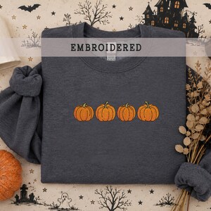 May include: A dark charcoal gray crewneck sweatshirt featuring an embroidered design of four orange pumpkins in a horizontal row across the center. The word EMBROIDERED is printed in black capital letters near the top of the garment.