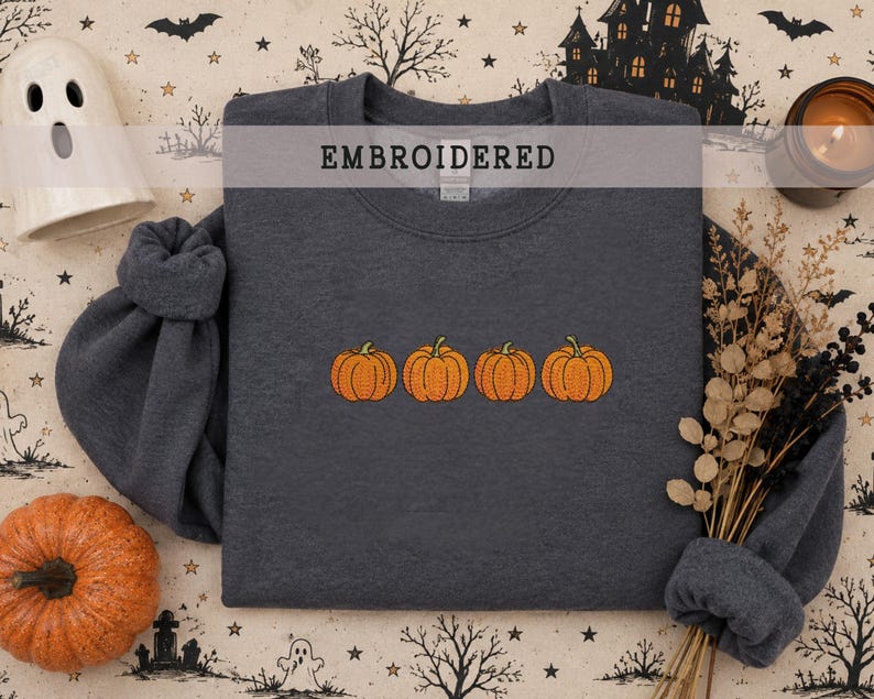 May include: A dark charcoal gray crewneck sweatshirt featuring an embroidered design of four orange pumpkins in a horizontal row across the center. The word EMBROIDERED is printed in black capital letters near the top of the garment.