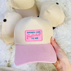 Peut inclure: Plusieurs casquettes de baseball crème et rose. L'une, tenue par une main, porte l'inscription "SOUNDS LIKE Bullshit TO ME" sur un patch rose. Les autres casquettes sont de couleur crème avec un bouton marron ou noir sur le dessus.