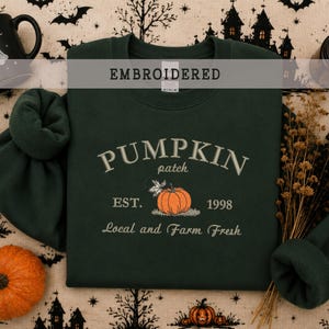 Op de afbeelding: Een donkergroene sweater met ronde hals en een geborduurd ontwerp met de tekst PUMPKIN patch EST. 1998 Local and Farm Fresh. De centrale afbeelding toont een oranje pompoen met een klein blaadje, alles geborduurd met lichtgekleurd garen.