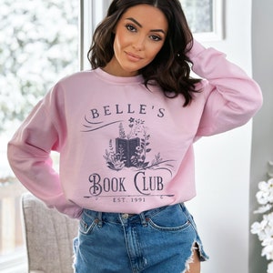 May include: A pink sweatshirt with the text "Belle's Book Club Est. 1991" and a graphic of an open book with flowers around it.
