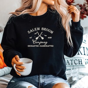 K&ouml;nnte beinhalten: Ein schwarzer Pullover mit einem wei&szlig;en Grafikdesign, auf dem "Salem Broom Company est. 1692 Enchanted + Handcrafted" steht. Die Grafik zeigt zwei gekreuzte Besen mit Flederm&auml;usen, die um sie herum fliegen.