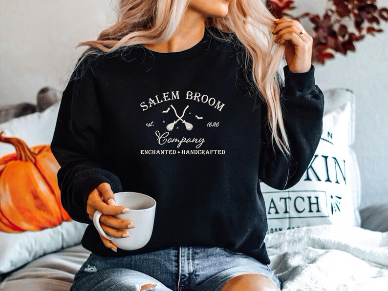 K&ouml;nnte beinhalten: Ein schwarzer Pullover mit einem wei&szlig;en Grafikdesign, auf dem "Salem Broom Company est. 1692 Enchanted + Handcrafted" steht. Die Grafik zeigt zwei gekreuzte Besen mit Flederm&auml;usen, die um sie herum fliegen.