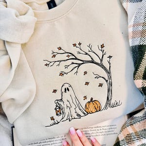 Pode incluir: Uma camisola de gola redonda bege com um design bordado de um fantasma a segurar uma lanterna, uma abóbora e uma árvore nua com folhas cor de laranja a cair. O bordado é feito com fios pretos, cor de laranja e castanhos na frente.