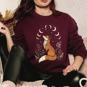 K&ouml;nnte beinhalten: Burgunderfarbenes Sweatshirt mit einer detaillierten Illustration eines Fuchses, der auf ein Mond- und Sternendesign blickt. Der Fuchs ist von floralen Elementen umgeben. Das Sweatshirt hat lange &Auml;rmel und einen Rundhalsausschnitt.