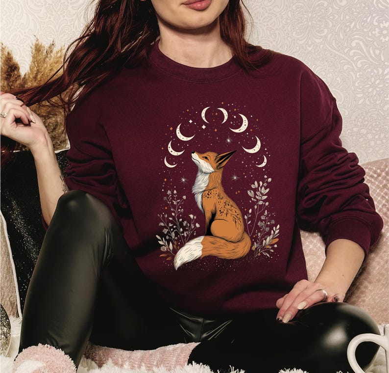 K&ouml;nnte beinhalten: Burgunderfarbenes Sweatshirt mit einer detaillierten Illustration eines Fuchses, der auf ein Mond- und Sternendesign blickt. Der Fuchs ist von floralen Elementen umgeben. Das Sweatshirt hat lange &Auml;rmel und einen Rundhalsausschnitt.