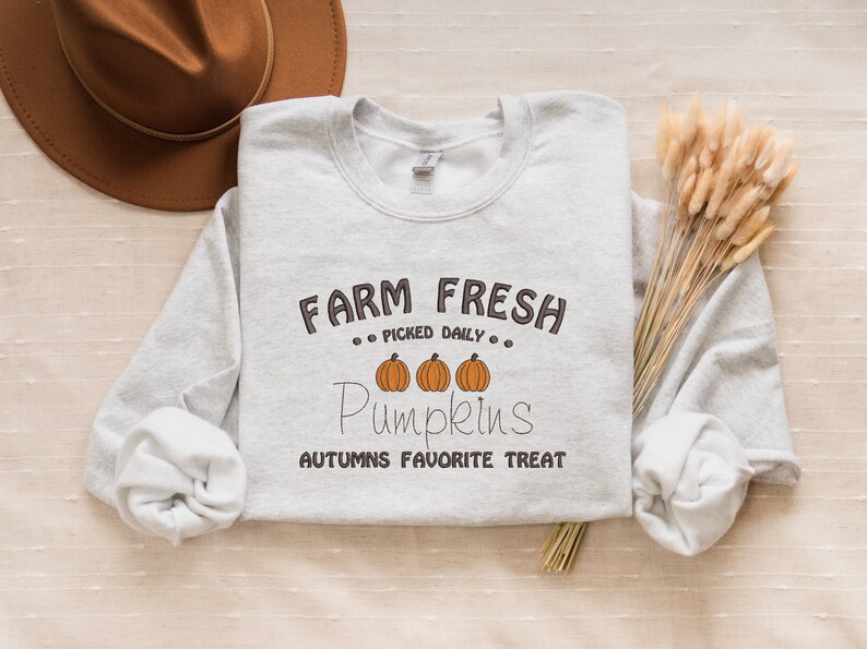 Op de afbeelding: Een lichtgrijs sweatshirt met de tekst "FARM FRESH, PICKED DAILY, Pumpkins, AUTUMNS FAVORITE TREAT" in bruin en oranje. Een bruine hoed en gedroogde tarwearen staan ook op de afbeelding.