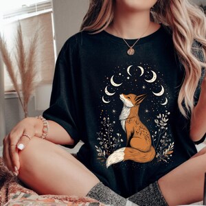 K&ouml;nnte beinhalten: Schwarzes T-Shirt mit einem Fuchs-Design in Orange und Wei&szlig;, das nach oben blickt, umgeben von Monden und Sternen. Das Design enth&auml;lt florale Elemente. Das T-Shirt hat einen l&auml;ssigen Stil.