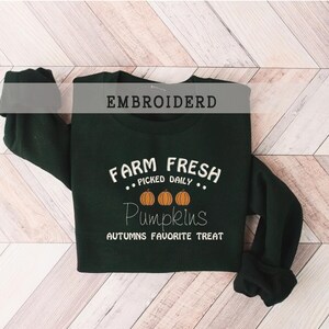 Op de afbeelding: Donkergroene sweatshirt met witte tekst "FARM FRESH PICKED DAILY Pumpkins AUTUMNS FAVORITE TREAT". Het ontwerp bevat drie oranje pompoenen. Het woord "EMBROIDERD" staat bovenaan de afbeelding.