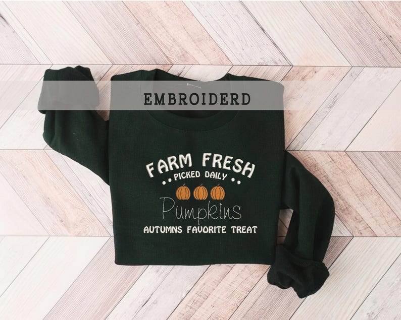 Op de afbeelding: Donkergroene sweatshirt met witte tekst "FARM FRESH PICKED DAILY Pumpkins AUTUMNS FAVORITE TREAT". Het ontwerp bevat drie oranje pompoenen. Het woord "EMBROIDERD" staat bovenaan de afbeelding.
