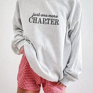 Pode incluir: Um moletom cinza claro com o texto "just one more CHAPTER" em preto, com um pequeno cora&ccedil;&atilde;o. A pessoa est&aacute; usando shorts xadrez vermelho e branco. A roupa &eacute; casual e confort&aacute;vel.
