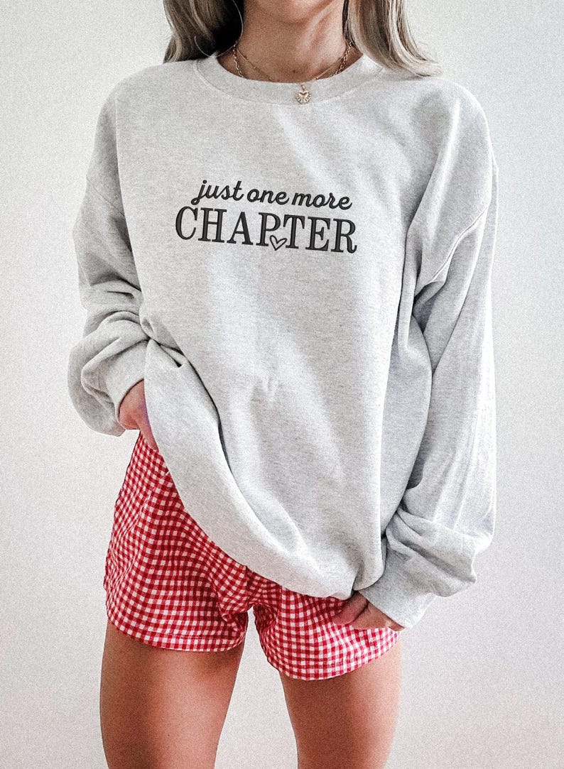 Pode incluir: Um moletom cinza claro com o texto "just one more CHAPTER" em preto, com um pequeno cora&ccedil;&atilde;o. A pessoa est&aacute; usando shorts xadrez vermelho e branco. A roupa &eacute; casual e confort&aacute;vel.