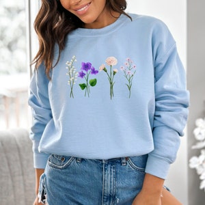 May include: A light blue sweatshirt with a floral design featuring white, purple, and pink flowers. The flowers are arranged in a row across the front of the sweatshirt.