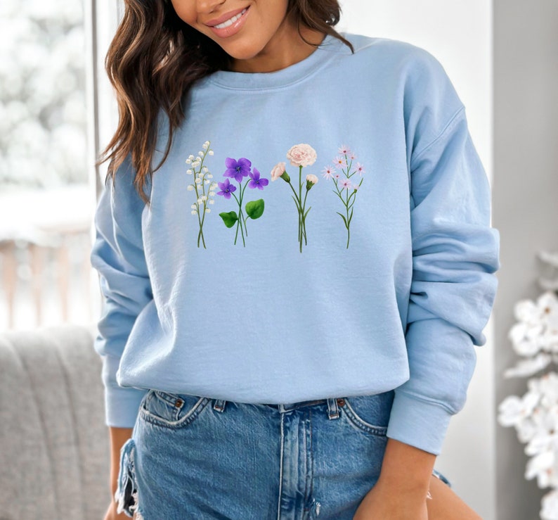 May include: A light blue sweatshirt with a floral design featuring white, purple, and pink flowers. The flowers are arranged in a row across the front of the sweatshirt.