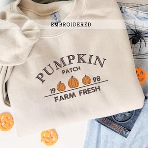 Könnte beinhalten: Beige besticktes Sweatshirt mit den Worten "PUMPKIN PATCH" und drei orangefarbenen Kürbissen. Der Text "1998 FARM FRESH" ist ebenfalls auf der Vorderseite gestickt. Das Wort "EMBROIDERED" steht über dem Design.