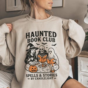 May include: Beige sweatshirt with a black graphic that reads "HAUNTED BOOK CLUB" above illustrations of a witch, a skeleton, and a ghost reading books. The phrase "SPELLS & STORIES - BY CANDLELIGHT -" is printed below the illustrations.