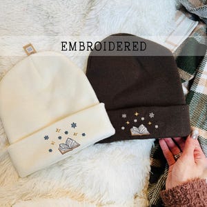May include: Two embroidered beanies. One is cream-colored, the other is dark brown. Both have a folded cuff with an embroidered design of an open book, stars, and snowflakes. The word "EMBROIDERED" is printed across the top.