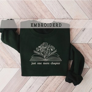 May include: A dark green sweatshirt with a white embroidered design of an open book with flowers and the text "just one more chapter". The word "EMBROIDERD" is printed at the top.