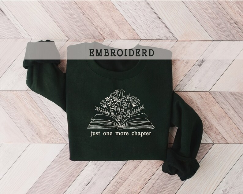 May include: A dark green sweatshirt with a white embroidered design of an open book with flowers and the text "just one more chapter". The word "EMBROIDERD" is printed at the top.