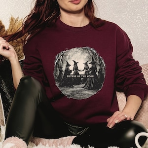 Può includere: Una felpa color bordeaux con una stampa grafica di quattro streghe in cerchio sotto una luna piena. Il testo "Sisters of the Moon" è stampato sotto le streghe.