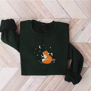 以下が含まれることがあります： かわいい刺繍の狐のデザインが施された濃い緑色のスウェットシャツ。狐は三日月と星を背景に上を見上げています。スウェットシャツはクルーネックで長袖です。