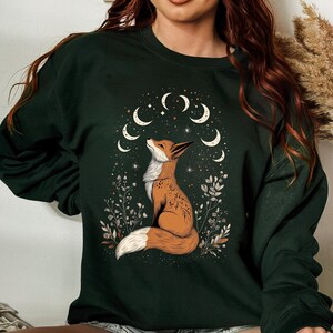 K&ouml;nnte beinhalten: Dunkelgr&uuml;nes Sweatshirt mit einer detaillierten Illustration eines Fuchses, der auf ein Mond- und Sternenmuster blickt. Der Fuchs ist braun und wei&szlig;, umgeben von floralen Elementen. Das Design ist mittig auf dem Sweatshirt platziert.